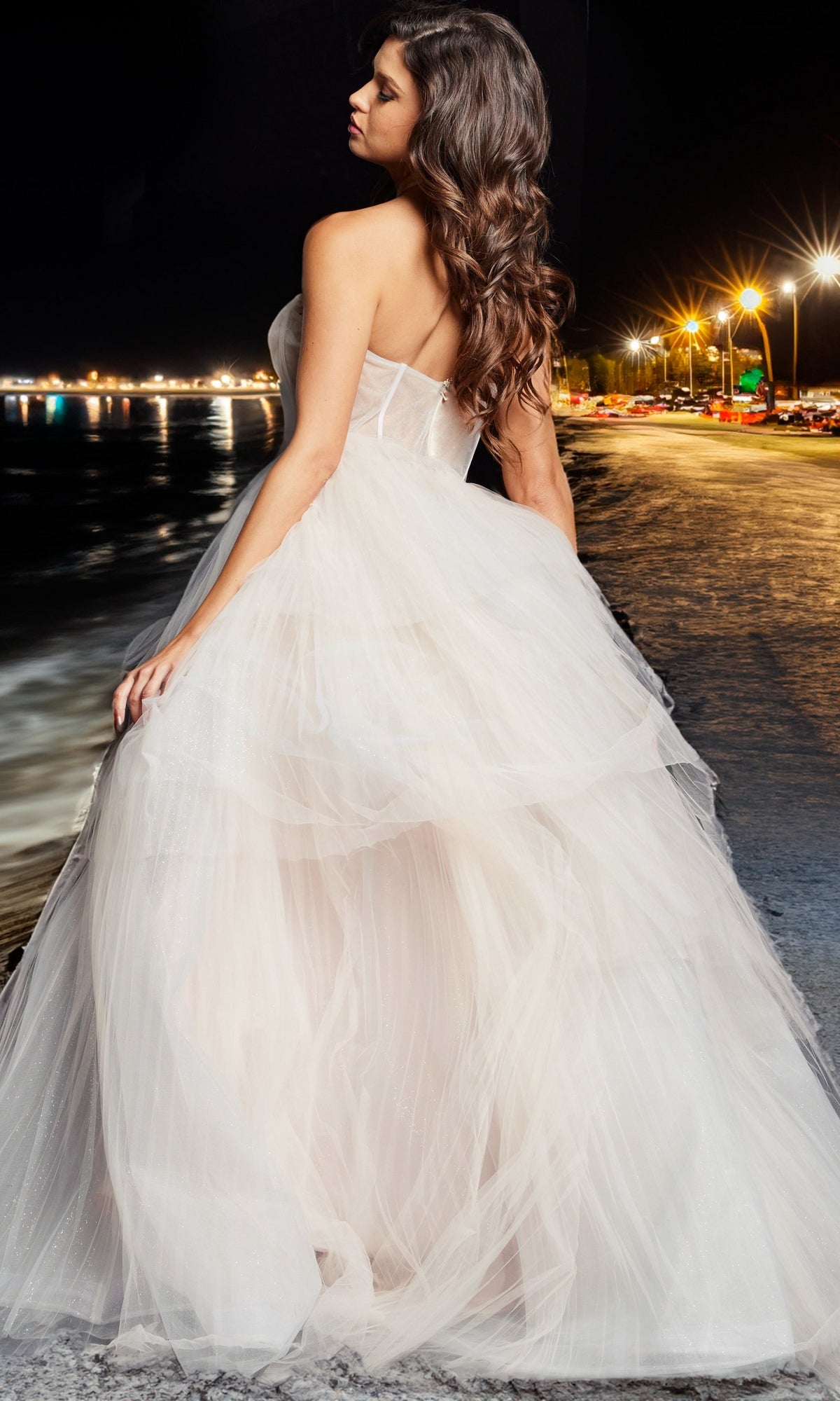 Wearing the Jovani Blush Pink Long Prom Ball Gown 38537 with a strapless corset-style sweetheart bodice, a woman stands on a waterfront at night, illuminated by shimmering streetlights and reflections.